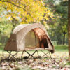 A raised cot tent combo, displayed erected on a leafy grass field, with trees in the background.