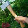 A hand holding a small hand axe with a wooden handle, along with its axe sheath displayed below, against a jungle background.