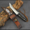 A Tanto fixed blade knife next to its leather sheath, lying against a small log.