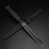 A black army dagger, displayed leaning against its knife sheath,