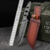A fixed blade tactical knife next to its leather sheath and a steel ruler, resting against a green ammunition crate.