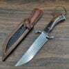 A damascus bowie knife and leather knife sheath laying on a wood floor.