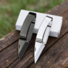 Two money clip knife models of differing colors displayed leaning against a small white box.