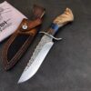 A wood handle fixed blade knife with a Damascus steel blade and its leather sheath lying on a concrete floor.