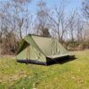An army green colored ridge tent, erected in a gass field, with trees in the background.