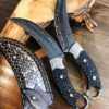 Two highly curved skinning knives and a leather sheath, lying against a wood tree branch.