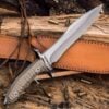 A large tactical belt knife and its leather sheath, resting against wood, with ammunition shells sporadically dispersed on the ground.