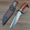 A Damascus steel tactical bowie knife with its leather sheath, lying on a wood floor.