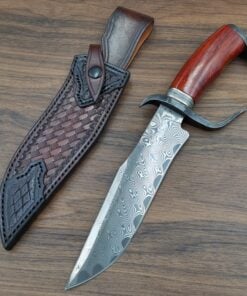 A Damascus steel tactical bowie knife with its leather sheath, lying on a wood floor.