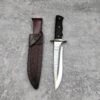 A large fixed blade combat knife with its leather sheath lying on a grey concrete floor.