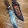 A Damascus steel western bowie knife nest to its leather sheath, lying on a wooden floor.