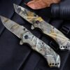 Two camouflage locking knives lying next to a leaf, against a black background.