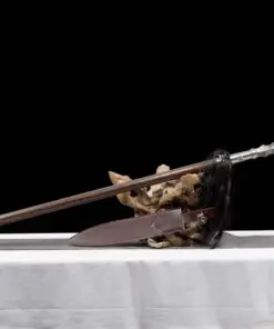 A battle ready spear leaning against a small wood log, with its scabbard below on the floor.