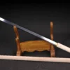A single edged, straight Japanese sword leaning against a wooden sword stand, with its wood scabbard displayed below.