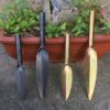 Four garden trowel tools of differing color and size, lined up against a planted pot.