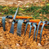 Five hand cultivator garden tools of differing designs, diplayed on earth ground.