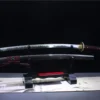A red and black sword Katana displayed next to its scabbard, on a small wooden stand.