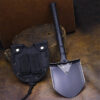 A tactical shovel displayed on the floor, leaning against a small log, with its black sheath next to it.