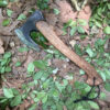 A bearded hatchet with a wooden handle, displayed lying on a leafy forest floor.
