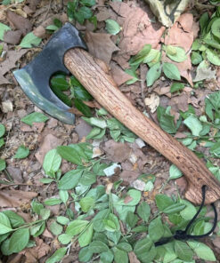 A bearded hatchet with a wooden handle, displayed lying on a leafy forest floor.