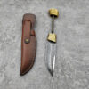 A bushcraft knife blank with a decorative brass bolster & pommel attached to the knife's tang, displayed next to its leather sheath on a cement floor.