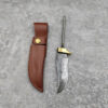 A Damascus steel knife blank with a brass bolster, displayed next to its leather sheath on a cement floor.