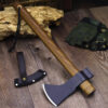A hand forged tomahawk axe with a wooden handle, displayed next to ist sheath, against a small wood log.
