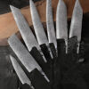 Seven kitchen knife blanks of varying design, displayed on a long wooden chopping board.