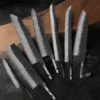 Seven Damascus steel kitchen knife blanks of differing design, displayed against a long wooden chopping board.