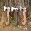 Three wood splitting hatchets with different color wooden handles, displayed side-by-side leaning against the base of a large tree.