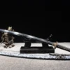 A Japanese Samurai Katana displayed on a small sword stand, with its decorative scabbard below on the floor.