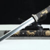 A ceremonial dagger with a straight, double edged blade, leaning against a small wooden stand, with its decorative sheath below on the floor.
