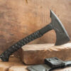 A tactical fighting tomahawk displayed standing balanced on a slab of wood, along with its axe scabbard below.