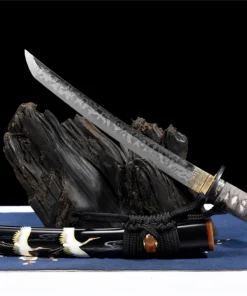 A Tanto Samurai knife displayed leaning against a small wood log, with its illustrated sheath below on the floor.