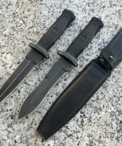 Two special forces knife models, displayed next to a sheath, on a stone-cement floor.