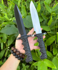 A mans hand holding two war knife models, against a leafy bush background.