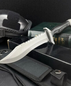 A combat survival knife displayed on a book, box and its sheath, with a black tactical helmet in the background.