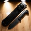 A black bushcraft survival knife, displayed nest to its sheath.