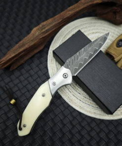 A Damascus folding pocket knife, displayed leaning against a small black box.