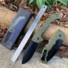 An extra large folding knife displayed next to a steel ruler & box, lying on a forest floor.