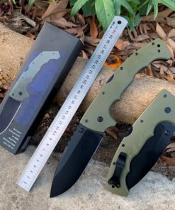 An extra large folding knife displayed next to a steel ruler & box, lying on a forest floor.